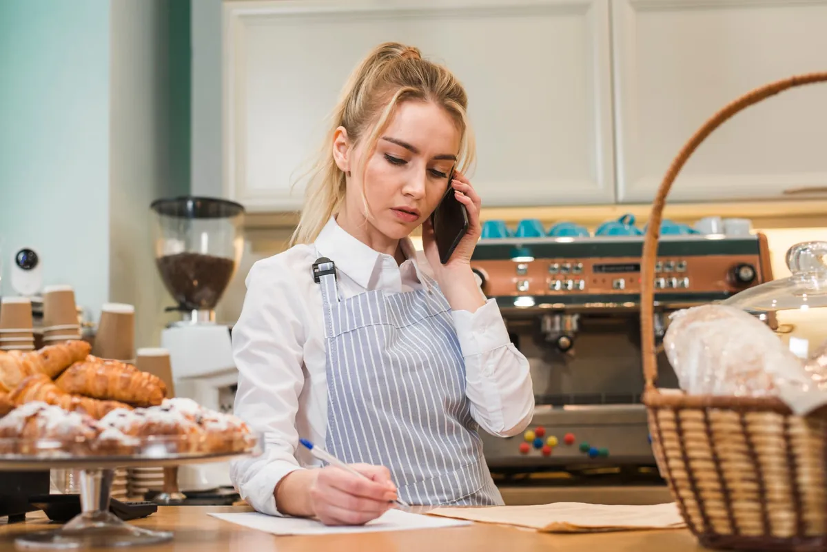 Dona de padaria anotando um pedido recebido pelo telefone, atrás do balcão